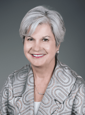Portrait of a smiling middle-aged woman with short, gray hair, wearing pearl earrings, a patterned blazer, and a necklace, against a gray background.