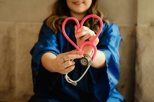 A woman in a blue jacket holding a pink stethoscope shaped like a heart, with the diaphragm of the stethoscope near her waist, smiling.